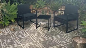 Un espace extérieur aménagé avec deux chaises noires et une petite table, le tout entouré d’une végétation luxuriante.
