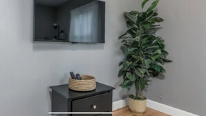 A room with a dark-colored nightstand, a woven basket with remote controls, a potted plant, and a wall-mounted mirror. - Cozy home in Bangor Maine (Bangor)