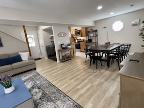 A modern living room with a sofa, a dining table with chairs, a kitchen area with cabinets and appliances, and a large window.