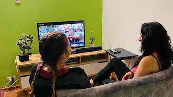 Una sala de estar con una pared verde, un televisor de pantalla plana y dos personas sentadas en un sofá viendo televisión.