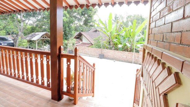 A covered balcony with a wooden railing, a brick wall, and a view of a residential area with houses and banana plants. - Homestay Nike Bed (Borobudur)