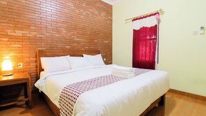 A neatly made bed with white linens and a patterned blanket, a wooden nightstand with a lamp, and a brick wall. - Homestay Nike Bed (Borobudur)