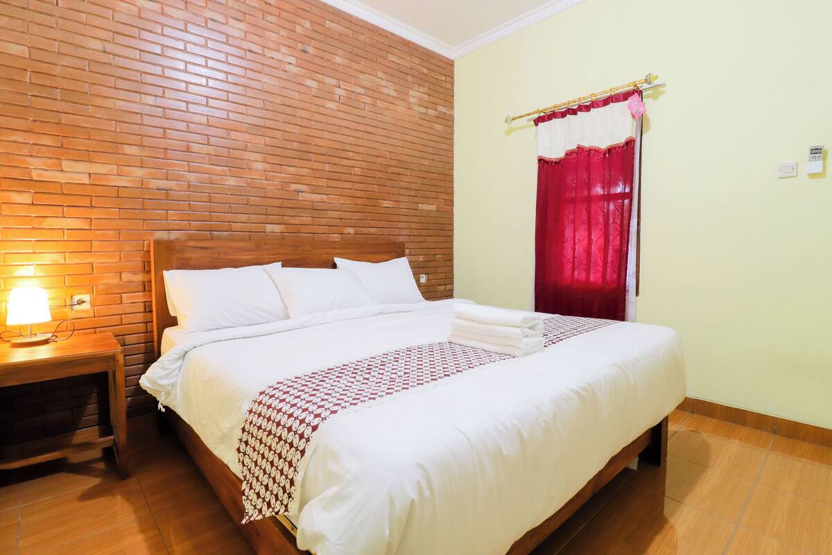 A neatly made bed with white linens and a patterned blanket, a wooden nightstand with a lamp, and a brick wall.