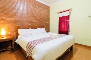 Una cama bien hecha con sábanas blancas y una manta estampada, una mesita de noche de madera con una lámpara, y una pared de ladrillo.