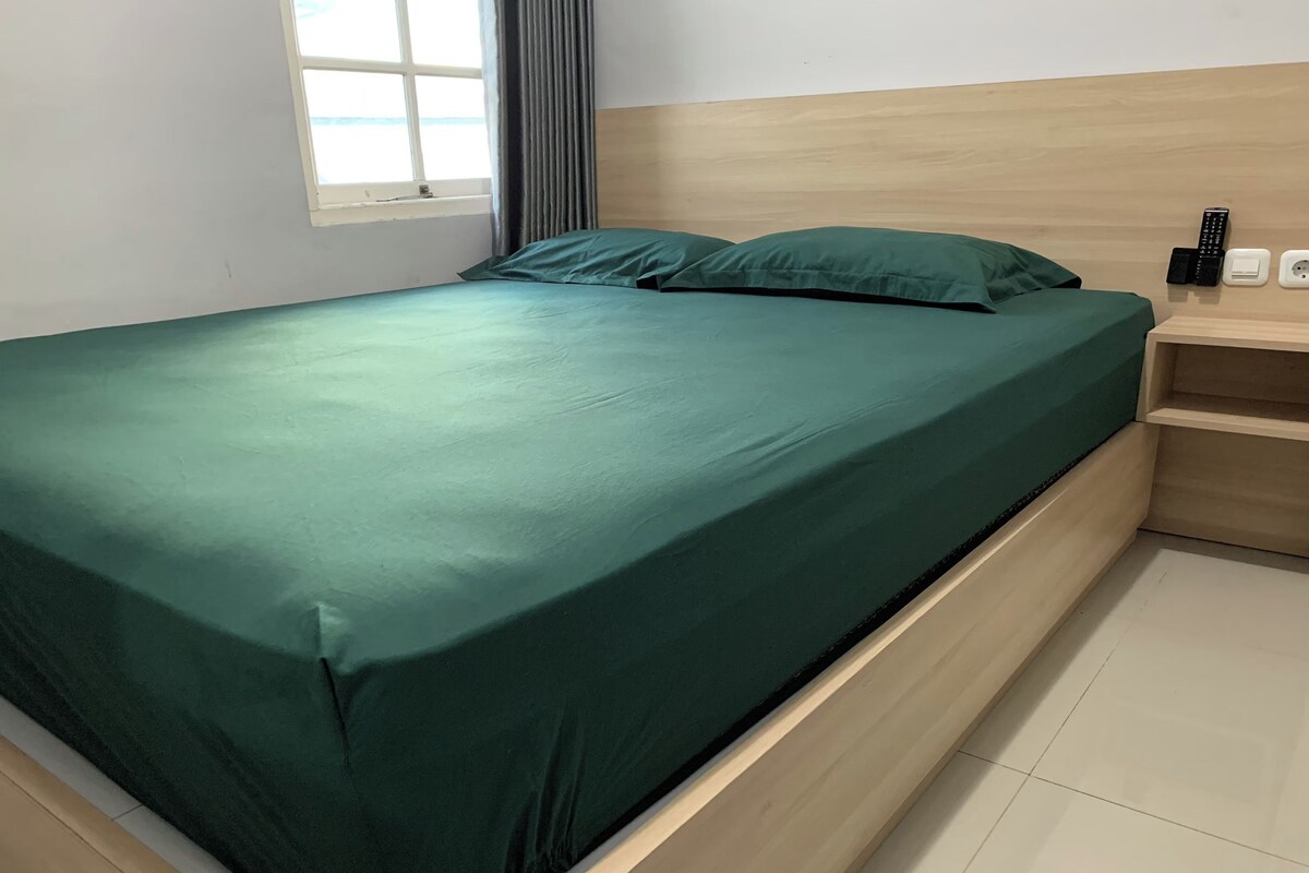 A neatly made bed with a green bedspread, a wooden headboard, a nightstand with a remote control, and a power outlet.