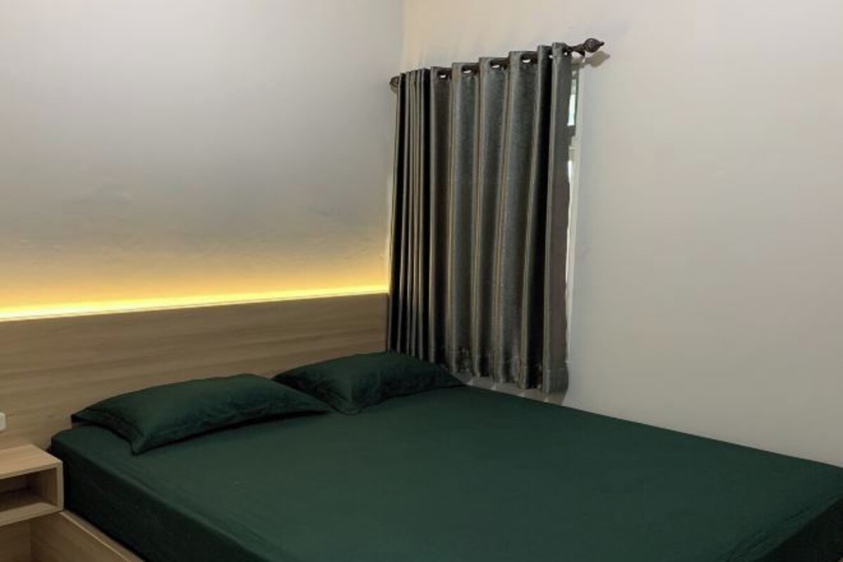 A single bed with a green bedspread and two pillows, a nightstand, and a window with dark curtains.