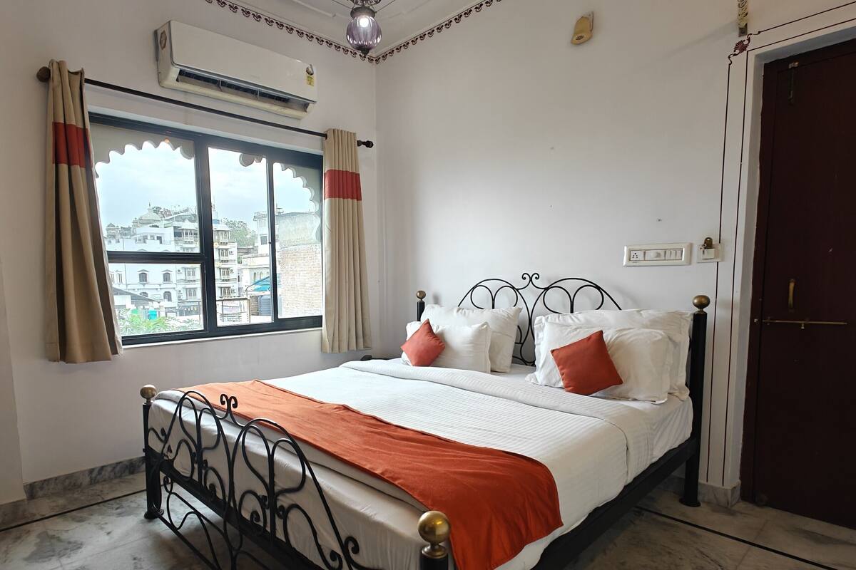 A bedroom with a bed, curtains, an air conditioning unit, a window with a view, a door, and a light fixture.