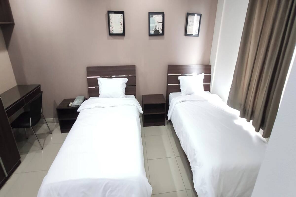 A hotel room with two single beds, a desk, a chair, and a small table with a phone.