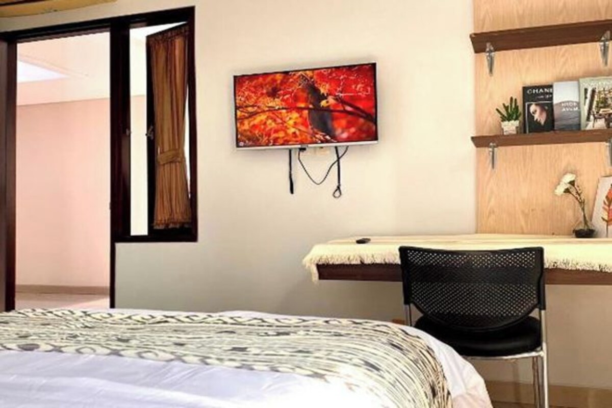 Una camera d'albergo con un letto, una scrivania con una sedia, una televisione a parete e una finestra con tende.