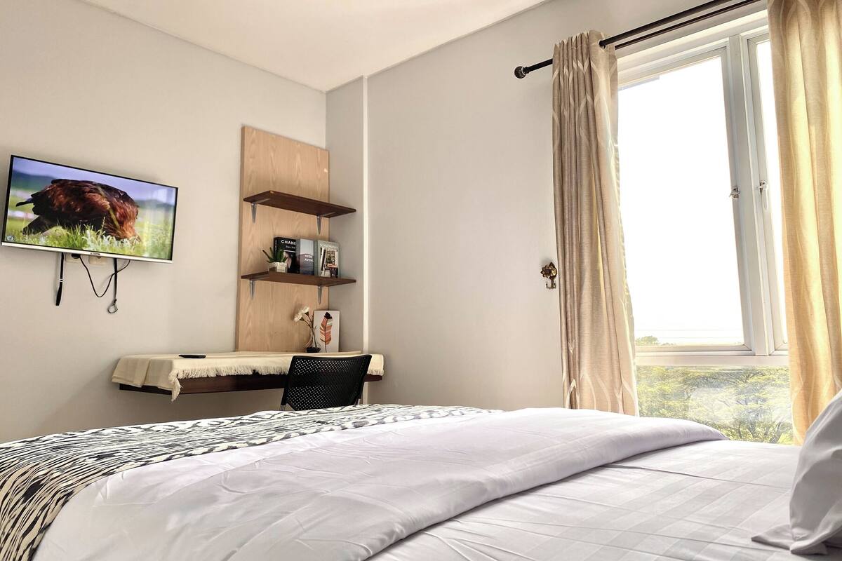 A bedroom with a bed, a TV mounted on the wall, a desk with a chair, and a window with curtains.