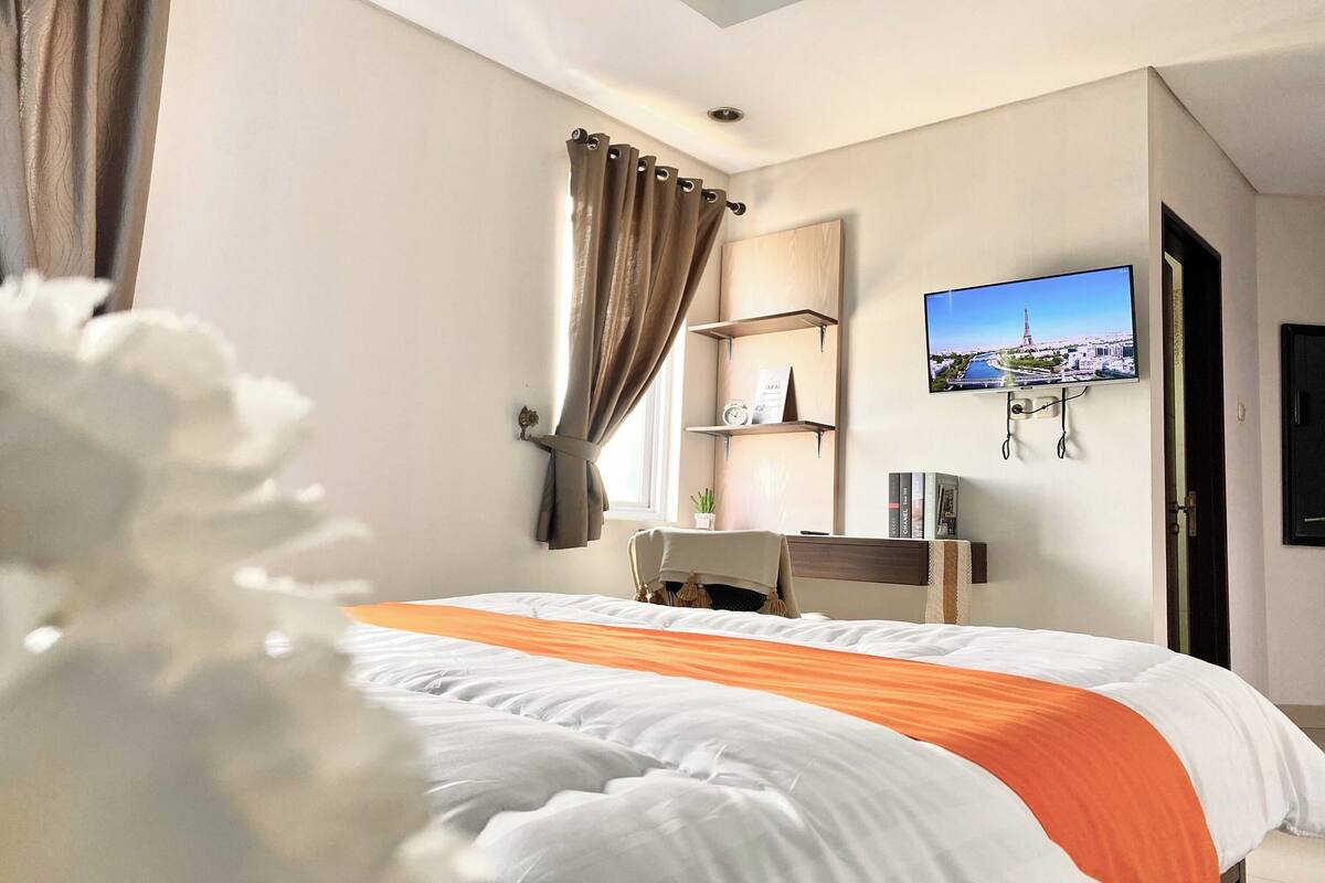 A hotel room with a bed, a TV mounted on the wall, a desk, and a window with curtains.