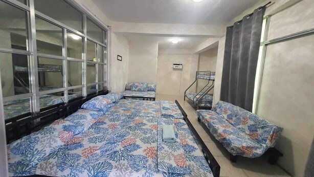 A room with a large bed, a smaller bed, and a sofa. There is a window with curtains, a ceiling light, and a wall-mounted air conditioning unit.