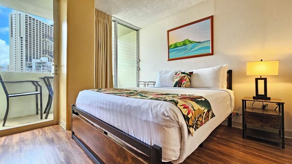 Quarto de hotel com cama, mesa de cabeceira com luminária, pintura na parede e vista para edifícios pela janela.