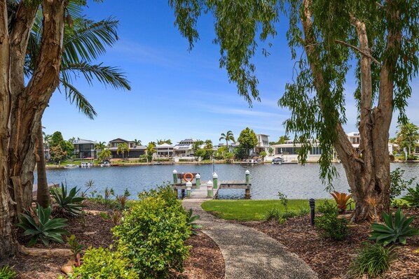 A lakeside view with a dock, surrounded by trees and greenery, leading to a waterfront property with houses in the background.