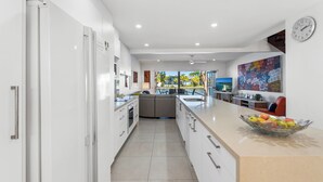 Une cuisine moderne avec des meubles blancs, un grand îlot central agrémenté d’un panier de fruits, un coin repas avec vue et une horloge au mur.