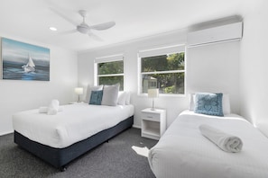 Two single beds with dark blue headboards and white bedding, ceiling fan, window with outdoor view, and wall-mounted air conditioning unit.