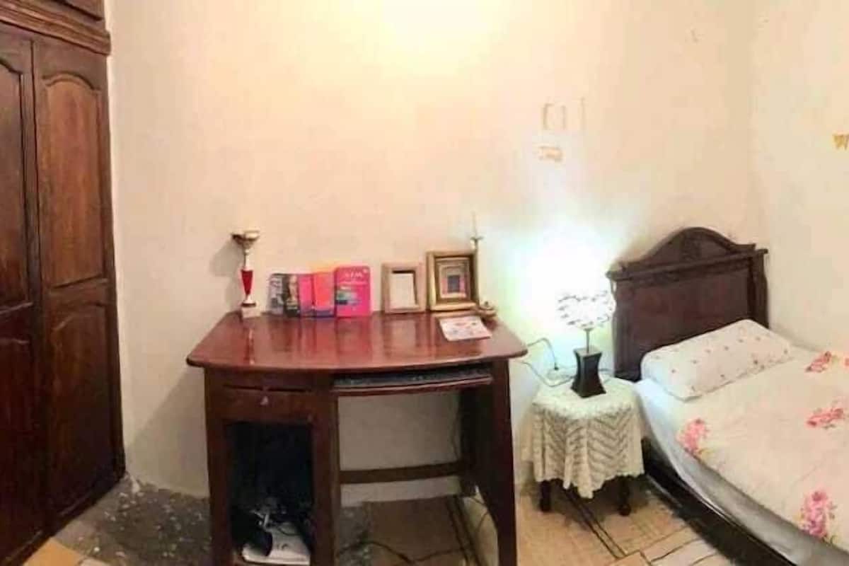 A small bedroom with a wooden desk, a bedside table with a lamp, a bed with floral bedding, and a wooden wardrobe.