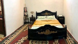 A bedroom with a black ornate bed, a patterned carpet, a nightstand, and a wall-mounted shelf with books. - Wohnung in Safi  (Safi)