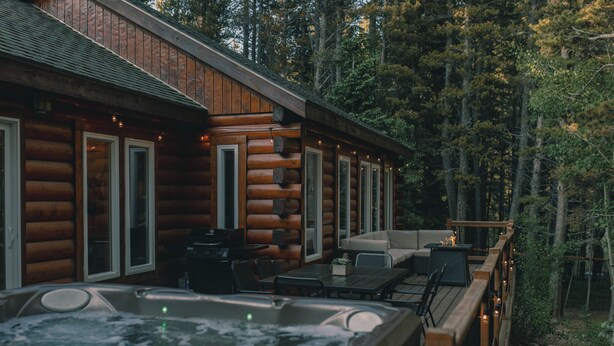 Una cabaña de troncos con jacuzzi, mobiliario exterior y una terraza con vistas a un bosque.