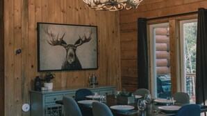 Interior de una cabaña de madera con una mesa de comedor para cuatro personas, una araña y un cuadro grande de cornamentas de ciervo en la pared.