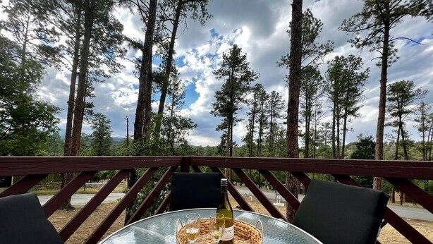 A glass table with a woven coaster, a bottle, and two glasses on a deck with a railing, surrounded by tall trees and a cloudy sky.