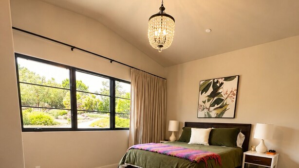 A bedroom with a bed, two bedside tables, a hanging light fixture, a window with curtains, and a wall-mounted painting.