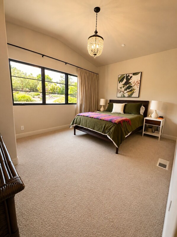 A bedroom with a bed, two bedside tables, a hanging light fixture, a window with curtains, and a wall-mounted painting.