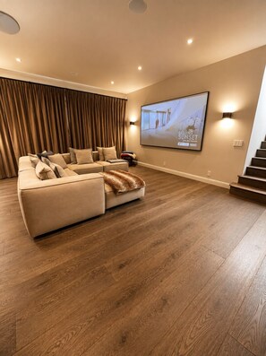 Sala de cinema com uma televisão de tela grande, sofá em formato de L e piso de madeira.