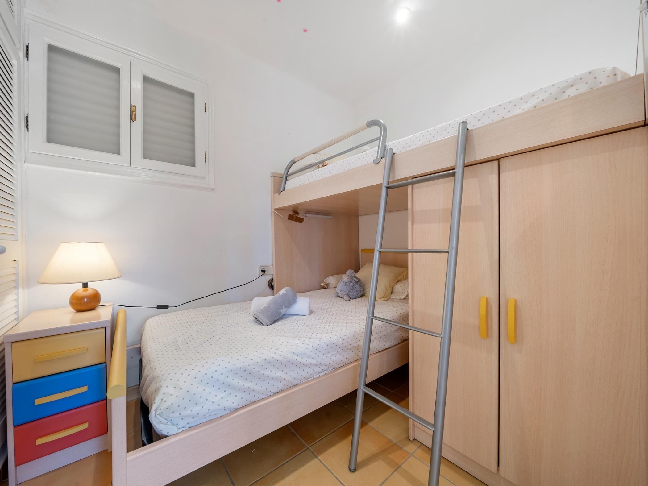 Una cama litera con escalera, una mesita de noche con lámpara y un armario de dos puertas.