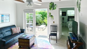 Un salon avec un canapé en cuir noir, une table basse, un ventilateur de plafond et une vue sur un jardin, visible à travers une porte vitrée.