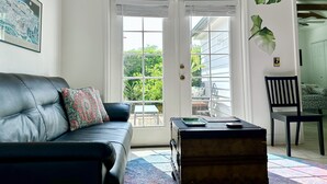 Un salon avec un canapé en cuir noir, un coussin décoratif à motifs, une table basse en bois et un tapis coloré. Il y a une grande fenêtre avec des stores, un tableau encadré au mur et une plante en pot.