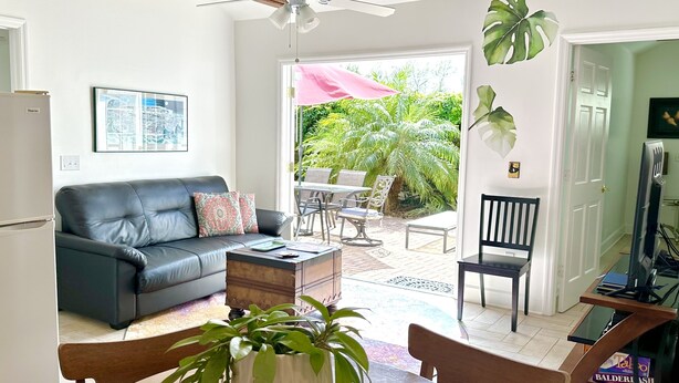 Un salon avec un canapé en cuir noir, une table basse blanche et une plante en pot. On y trouve également un ventilateur de plafond, un téléviseur et une porte donnant sur un balcon aménagé avec du mobilier d’extérieur.