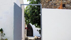 A narrow pathway leading to a dark wooden door, flanked by white walls and a stone wall. - The Three Sisters House (Kardamena)