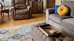 A living room with a brown leather armchair, a grey sofa with a yellow cushion, a wooden side table, a bookshelf, and a patterned rug. - Cozy Cottage in Downtown Redmond (Redmond)