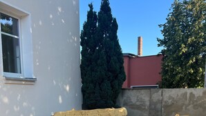 Une table de pique-nique en bois est placée sur une terrasse dallée, avec un bâtiment blanc et un arbre en arrière-plan.