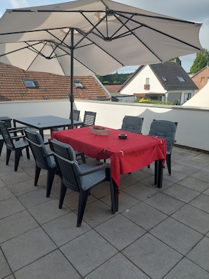 Un patio al aire libre con una mesa cubierta por un mantel rojo, rodeada de sillas, bajo un gran toldo.