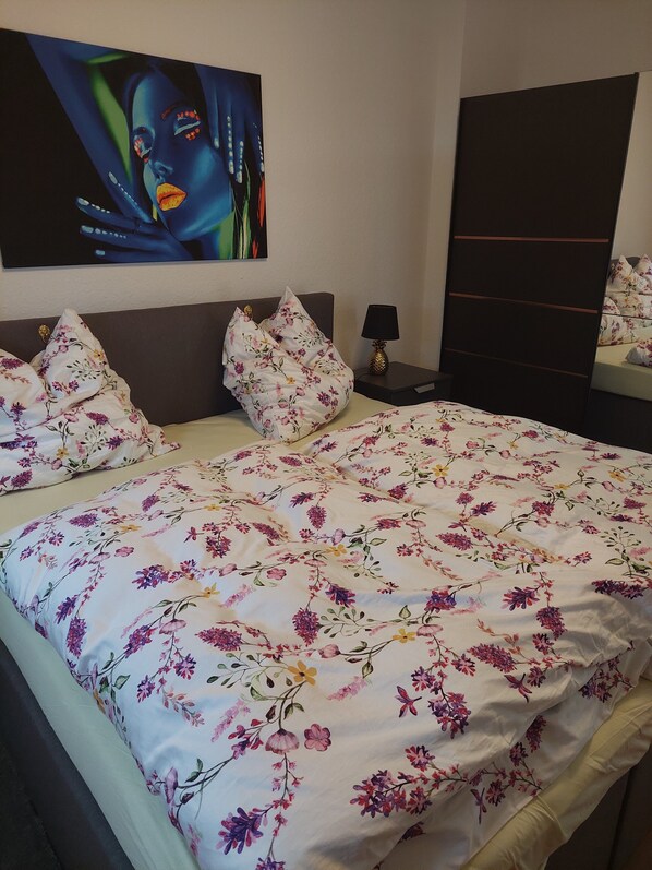 Une chambre à coucher avec un lit, une literie à motifs floraux, une table de chevet, une lampe, un tableau accroché au mur et un miroir.