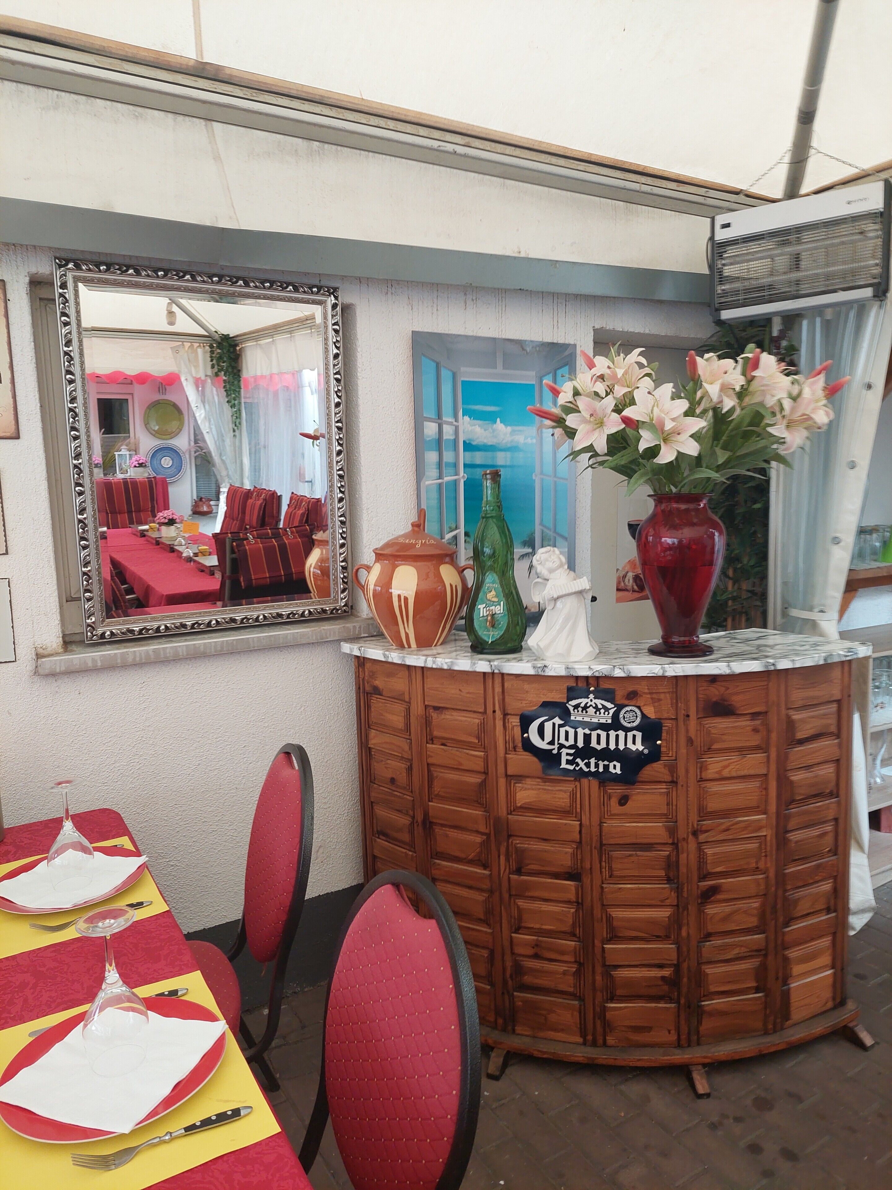 Un coin bar avec un miroir, un comptoir en bois, une enseigne indiquant « Corona Extra », un vase avec des fleurs et une table recouverte d’une nappe rouge, ainsi que des chaises.