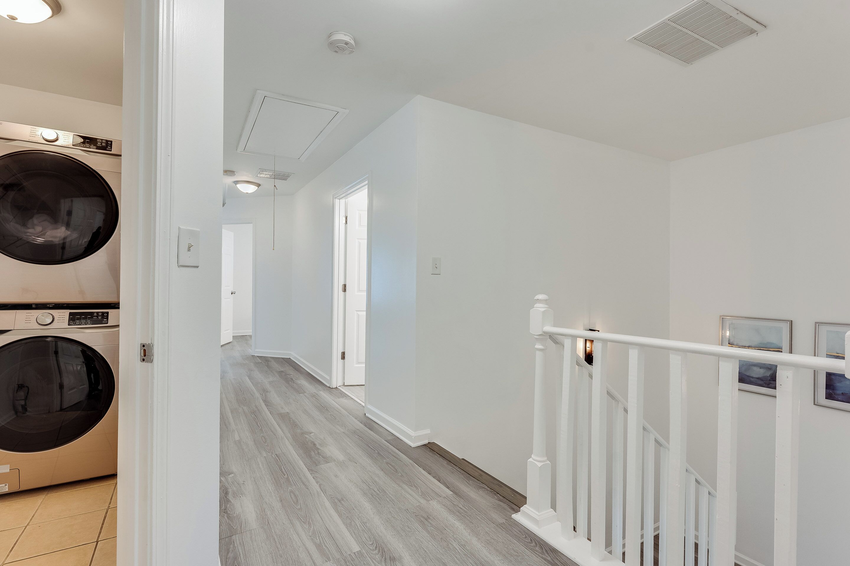 A laundry room with a front-loading washer and dryer, a hallway with a wooden floor, and a staircase with white railings.