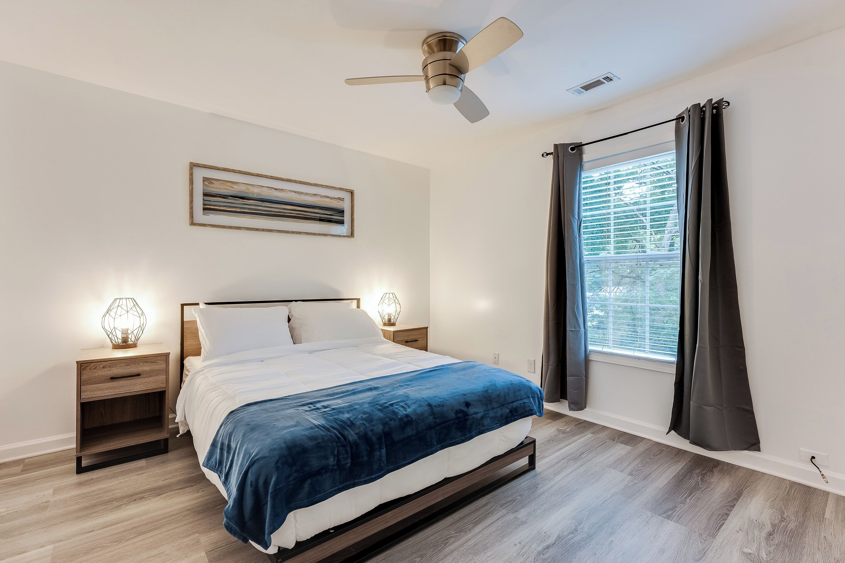 A bedroom with a bed, bedside tables, a ceiling fan, a window with curtains, and a wall-mounted artwork.
