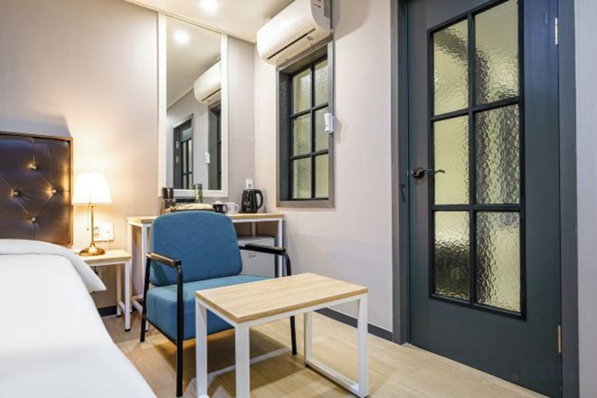 A hotel room with a bed, a desk with a chair, a small table, a mirror, and a window with frosted glass.