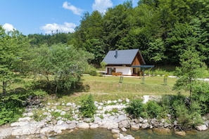 A cabin with a sloped roof, surrounded by a natural landscape with a stream and dense trees.