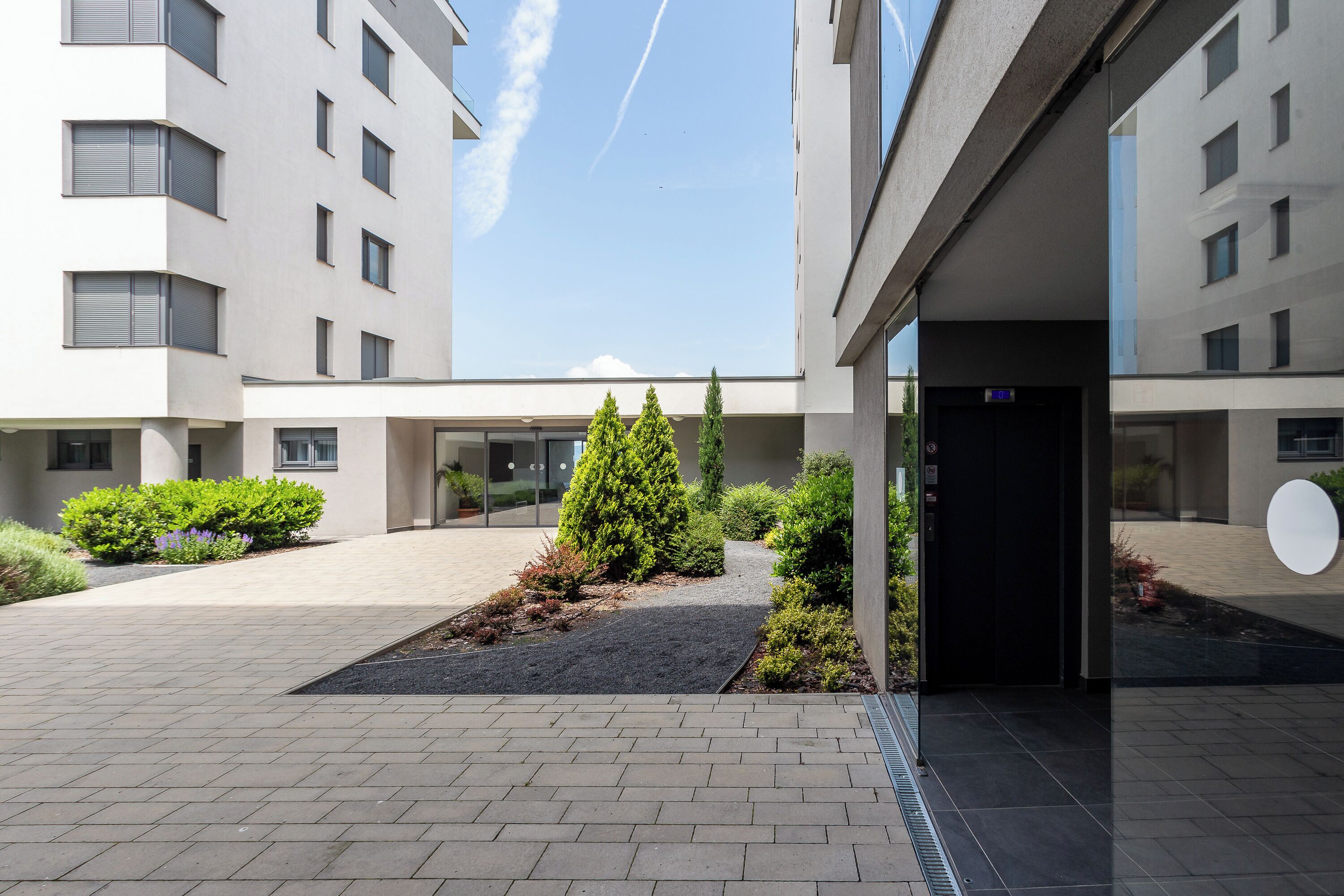 Un edificio moderno con un patio empedrado, un pequeño jardín con arbustos y un árbol, y una puerta negra con un letrero que dice 'EXIT'.