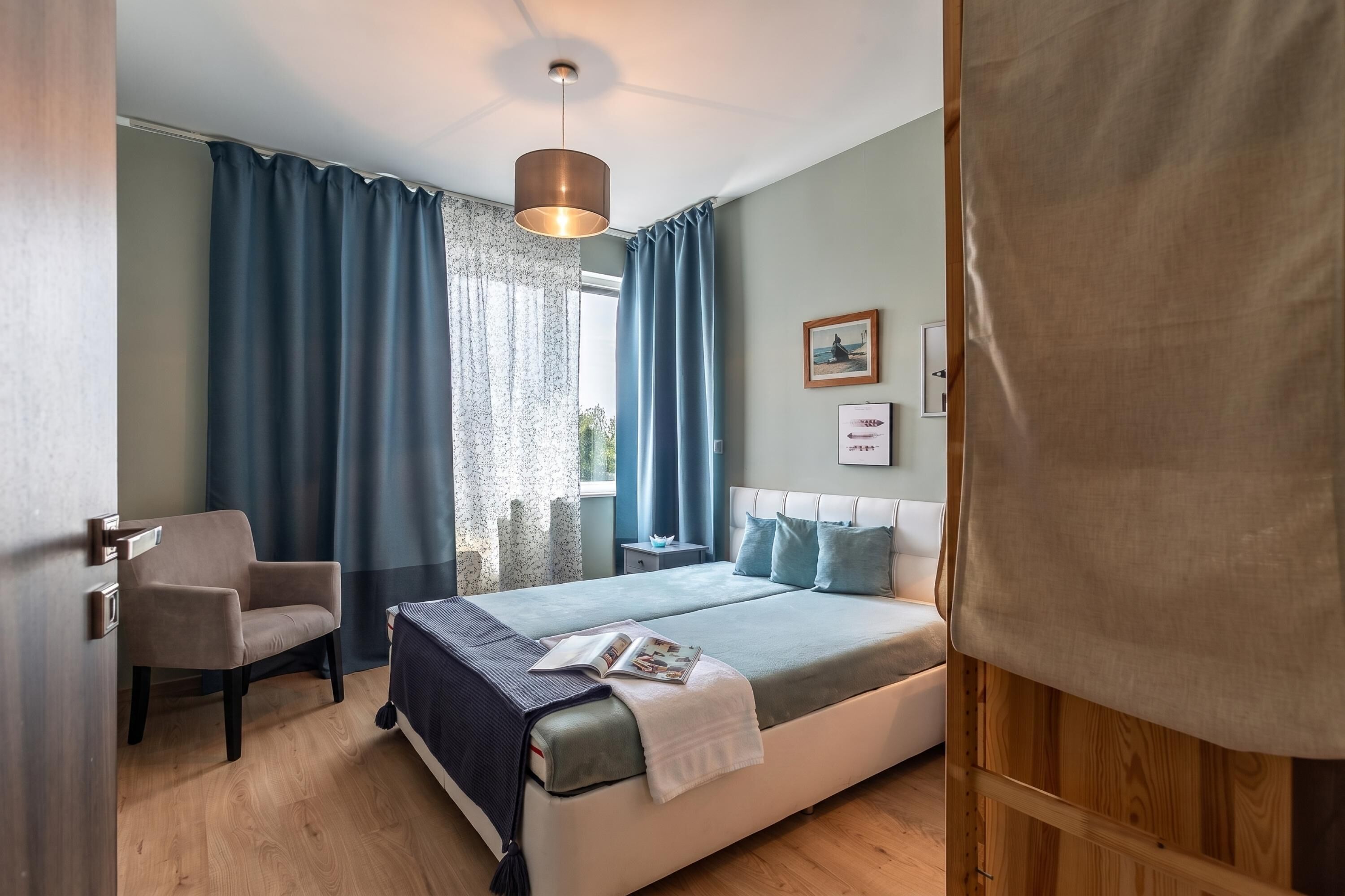 Een slaapkamer met een bed, een stoel, een nachtkastje, een lamp en gordijnen.