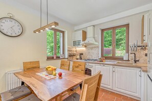 Une cuisine avec des meubles en bois, une table à manger avec des chaises, une horloge au mur et une fenêtre donnant sur un paysage verdoyant.