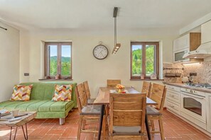 Un coin repas avec une table et des chaises en bois, un canapé vert et une cuisine dotée de meubles blancs et d’une cuisinière.