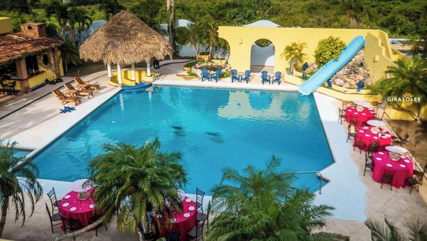 A large swimming pool with lounge chairs and tables, surrounded by palm trees and a thatched-roof structure. - Hacienda Girasoles Tampico (Pueblo Viejo)