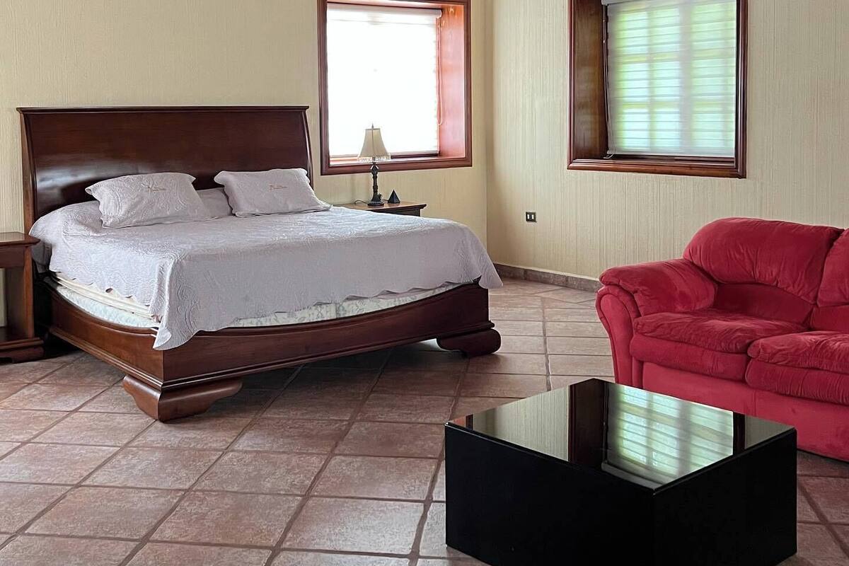 Une chambre avec un plafond en bois, un lit, deux fenêtres, une chaise rouge et une table noire.
