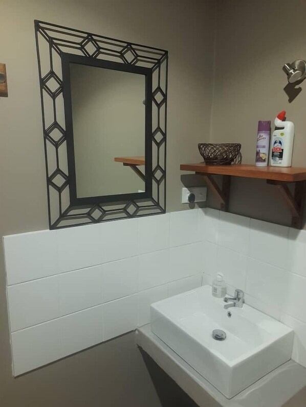 A bathroom with a white sink, a mirror with a geometric frame, and a shelf with toiletries. - Secured Apartments (Ndola)