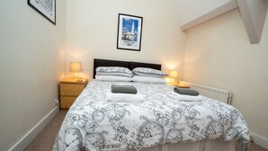 Een net opgemaakt bed met een sprei met klokkenprint, twee bedlampjes en een lijst met een foto van een vuurtoren aan de muur.